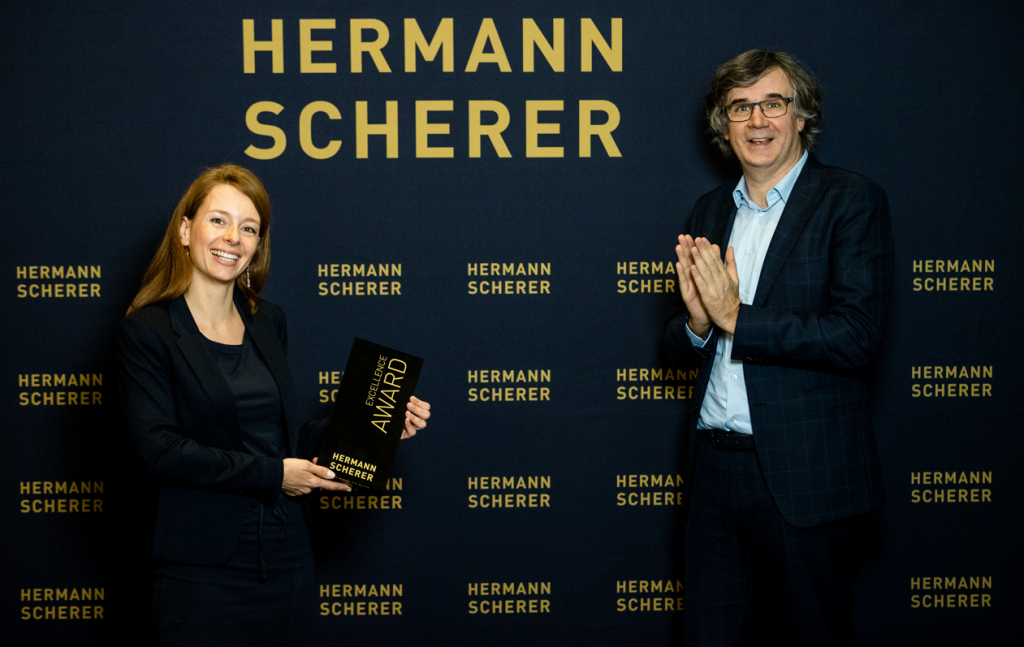 Anja Graffius hält einen Excellence Award in den Händen, während Sie vor einer Fotowand posiert und mit Hermann Scherer abgelichtet wird